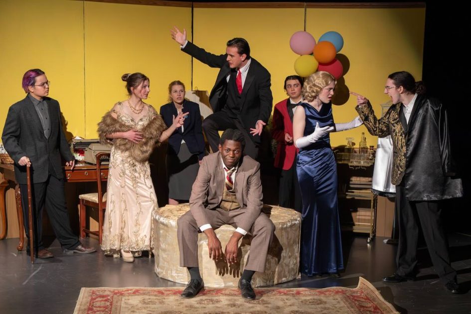 Skyline Light Up the Sky Production photo - Cast of students dressed in suits and formal dresses in front of a yellow background. central man seated on an ottoman looking defeated while a man stands behind him on the ottoman gesturing wildly. On the right a man points his finger in the face of a woman.
