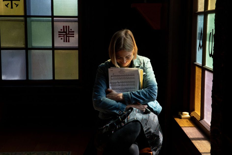 Pianist Anne Lovering Rounds sits in the dark lit through stained glass windows cradling sheet music