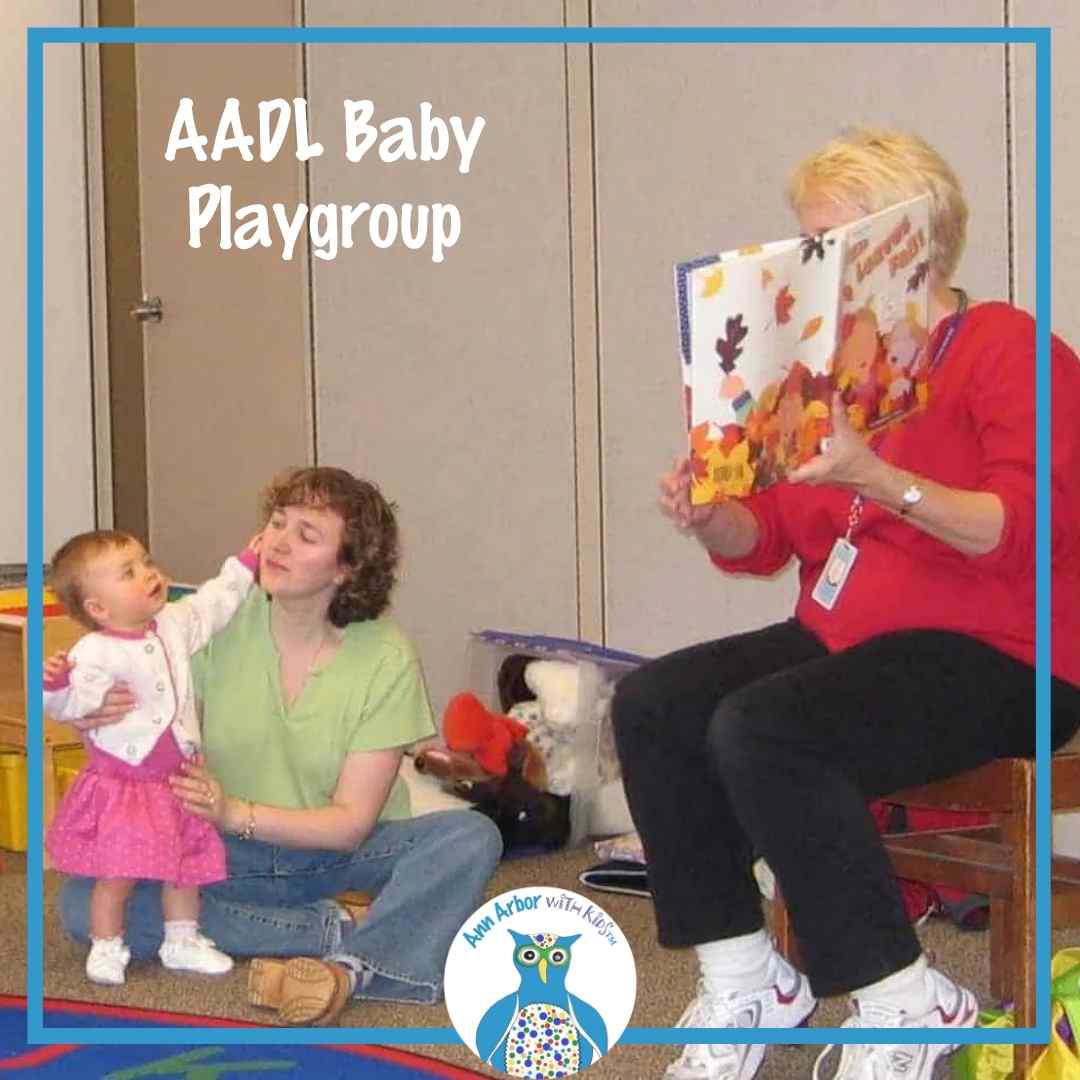 AADL Baby Playgroup - a baby standing supported by mom watches a storyteller holding a book