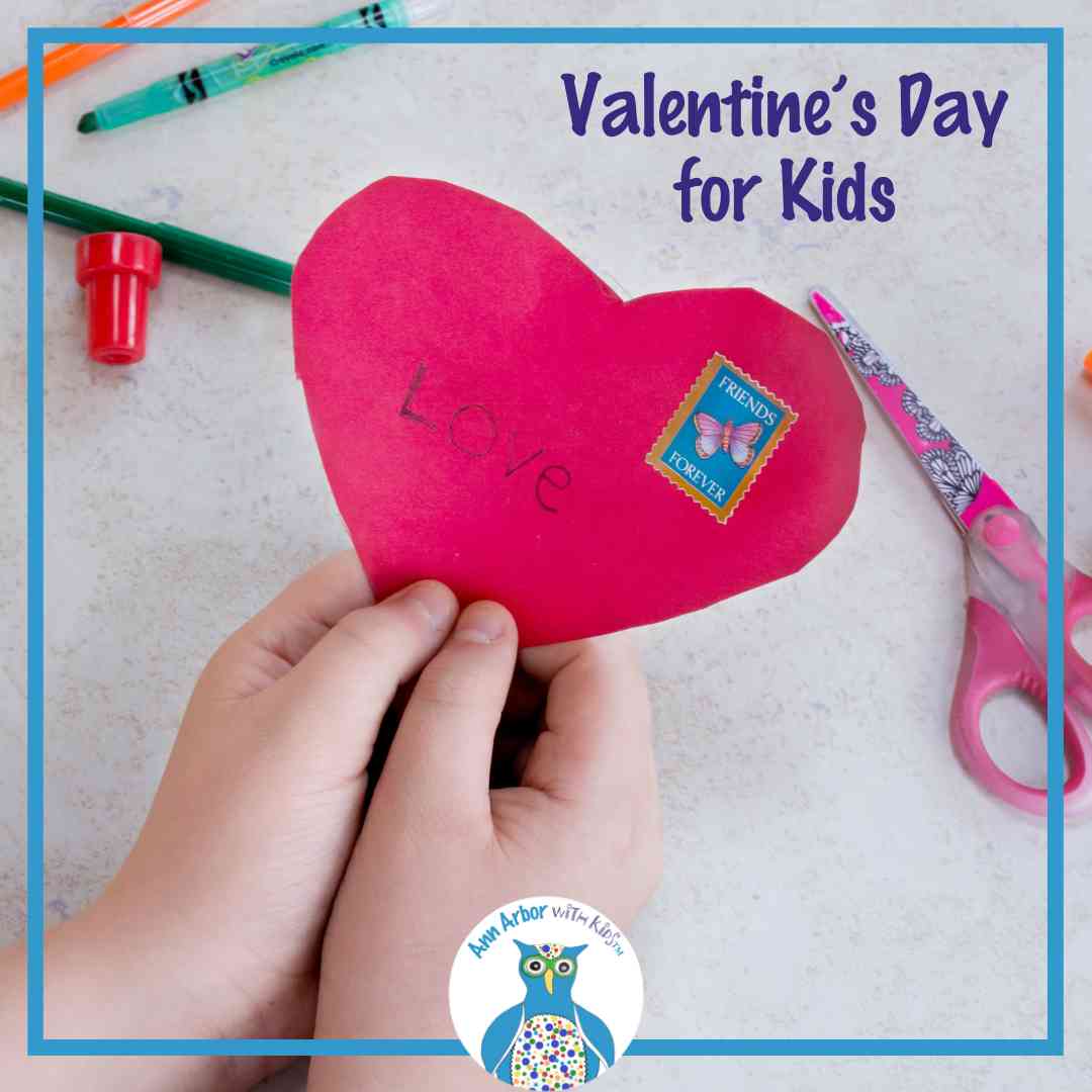 Valentine's Day for Kids - a child holds a construction paper heart that says LOVE and has a sticker over a table with scissors and other craft supplies