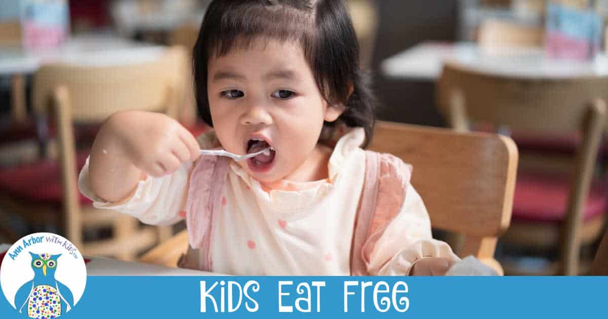 Ann Arbor Kids Eat Free - a little girl sitting in a high chair at a restaurant feeding herself with a spoon
