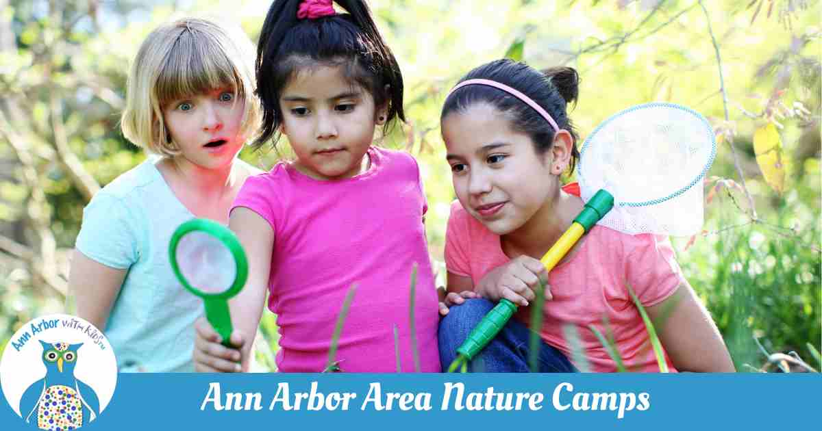 Ann Arbor Nature Camps - 3 girls use a magnifying glass to look in a field, one carries a net