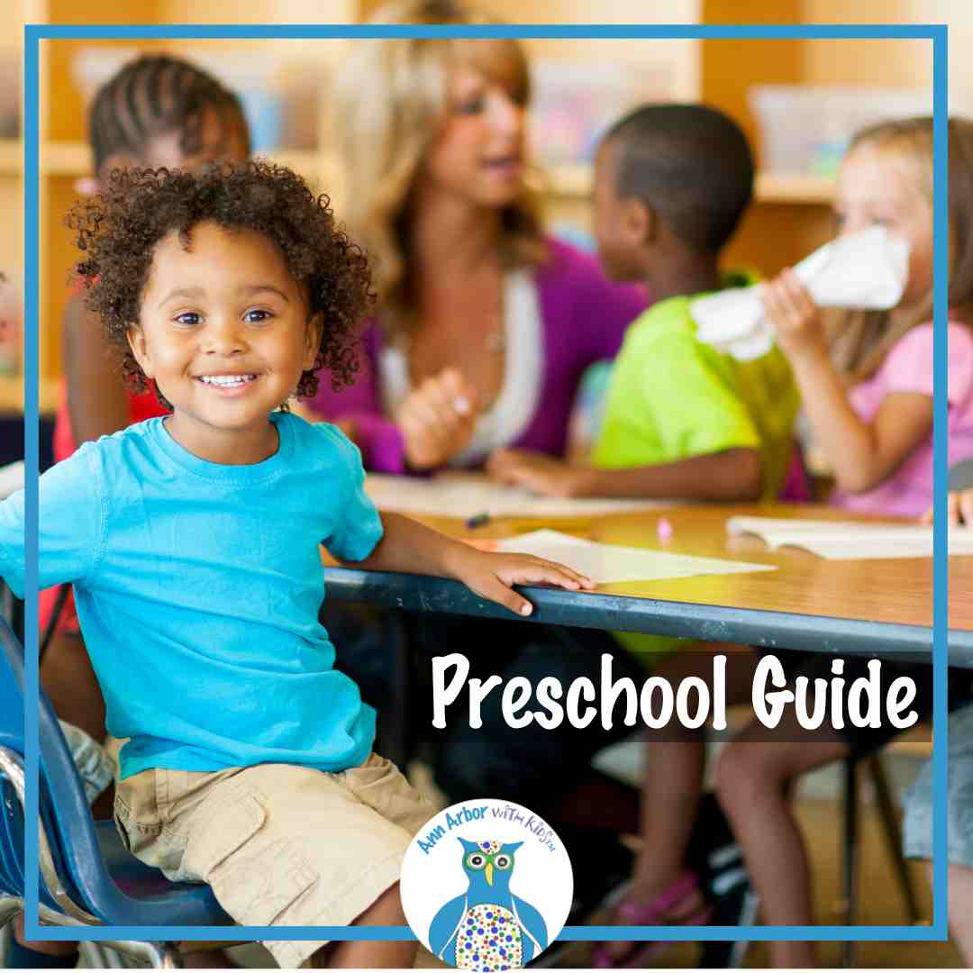 Ann Arbor Preschool Guide - a preschool class. A boy sits at table and looks at camera. Out of focus behind him is a teacher squatting down to talk with other classmates at the table