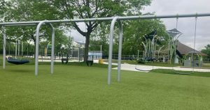 Bicentennial Park - 2026 New Additions - Swings including saucer, baby, and regular, play structure with merry go rounds, and splash pad in the distance