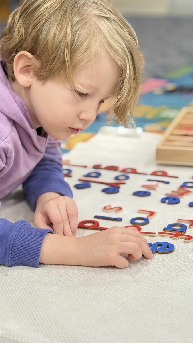 Daycroft School - a preschooler lays on the floor working with letters to form words - vowels are blue and consonants are red