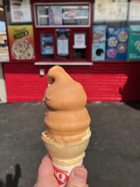 Free Cone Day - a caramel dipped Dairy Queen cone held out in front of camera with a closed walk up window behind.. Building is red and windows are covered in posters promoting blizzards and other items.