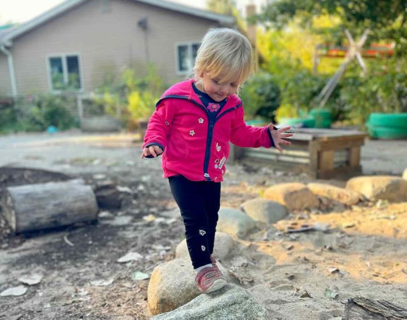 Green Apple Garden Lower School - A toddler balances on rocks around a sand play area