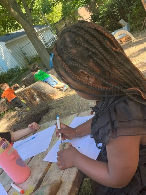 Child sits at a picnic table using markers to write on a chunk of wood. Papers on table. Other kids play over stumps in background