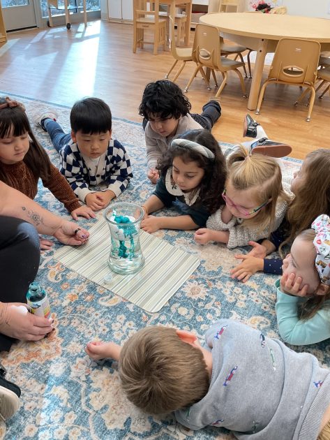 Ivybrook Academy - Kids lay on carpet watching a science experiment of water in a clear vase with blue liquid swirled in.