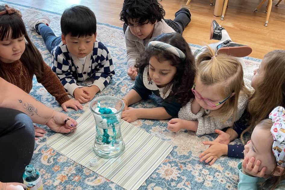 Ivybrook Academy - Kids lay on carpet watching a science experiment of water in a clear vase with blue liquid swirled in.