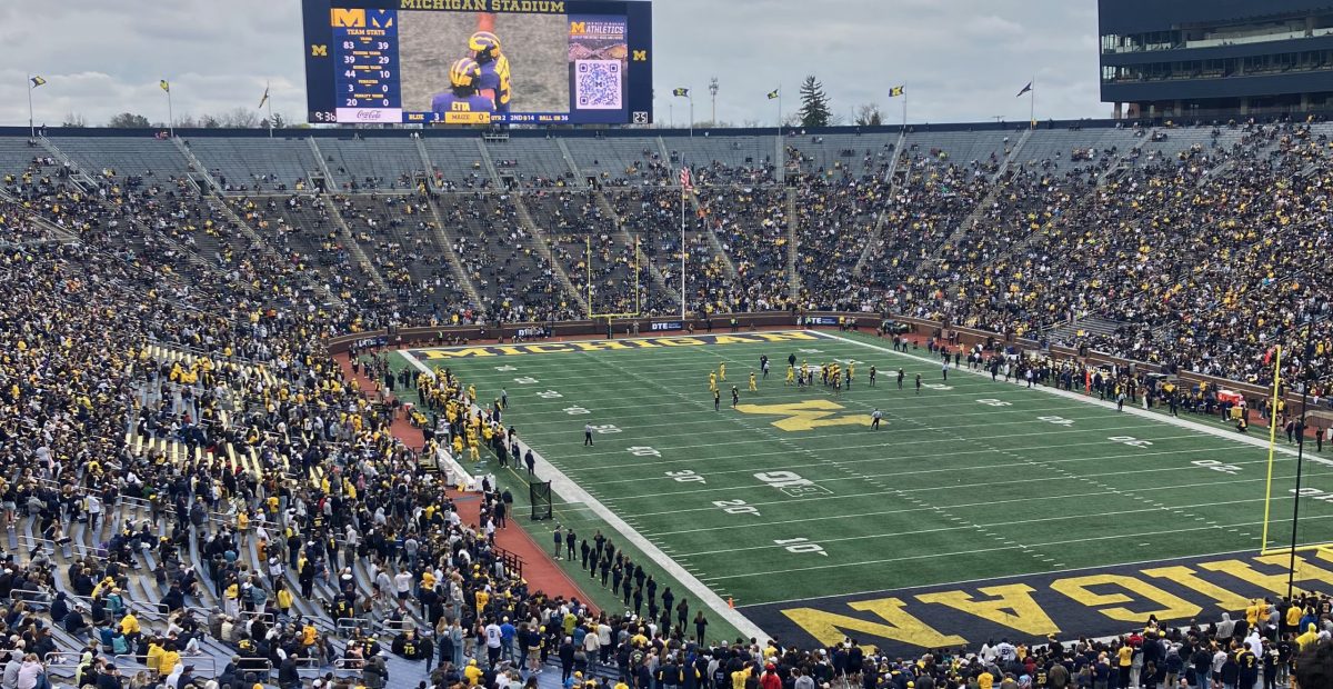 Michigan Football Spring Game - photo taken from north-east corner seating. Stadium is half-full for the spring game. Scoreboard has closeup of player with several players on the field around the 35 yard line - some players in blue uniforms and some in maize