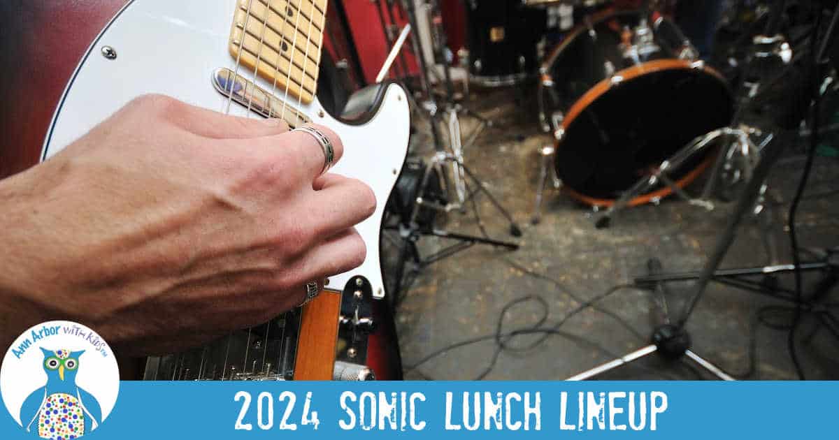 2024 Sonic Lunch Lineup - Closeup of a hand strumming an electric guitar with drums int he background.