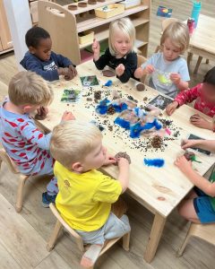 Green Apple Garden Playschool - Upper School - Preschoolers sit indoors at a table playing with clay, pine cones, and other natural materials