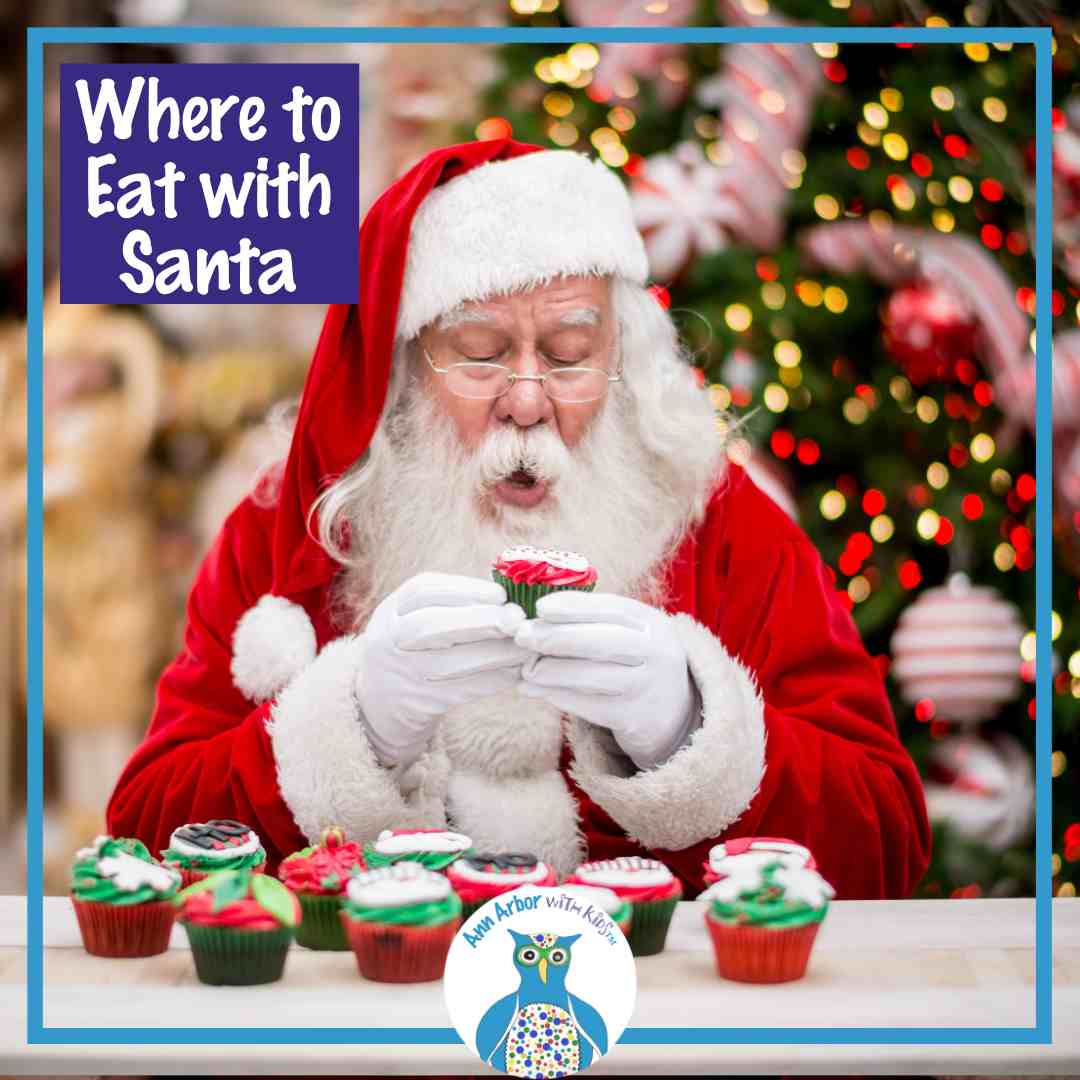 Where to Eat with Santa -Santa sits at table topped with festive cupcakes while he holds one . Decorated Tree is behind him